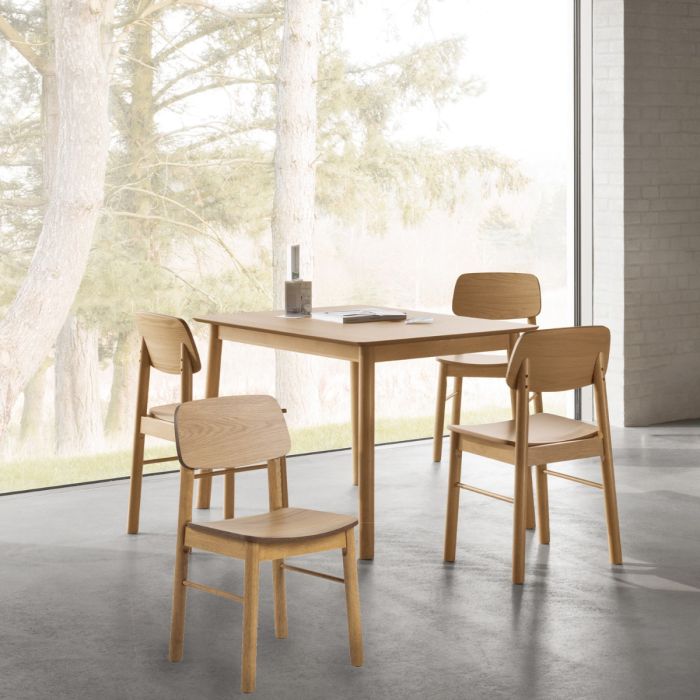 Wooden dining table with four chairs in a room with large windows showing trees outside.