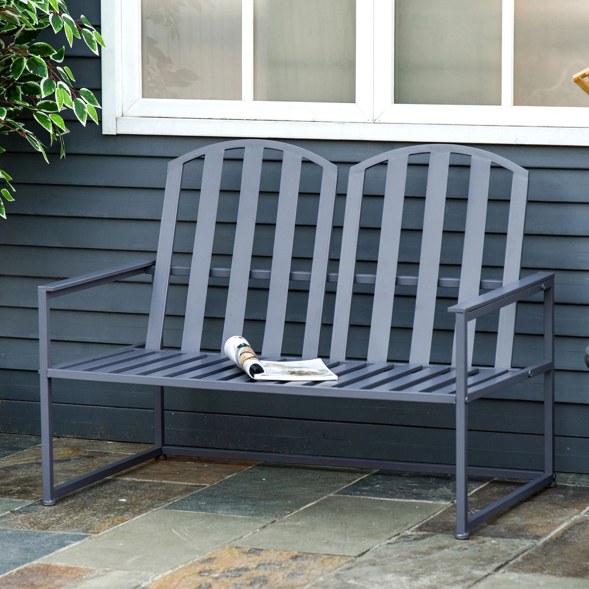 Gray metal bench on a stone patio with a gray wall and window in the background